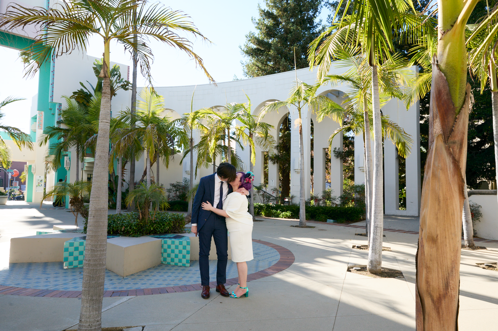 Couple sharing a romantic moment among palm trees and white arches at a sunny outdoor venue.