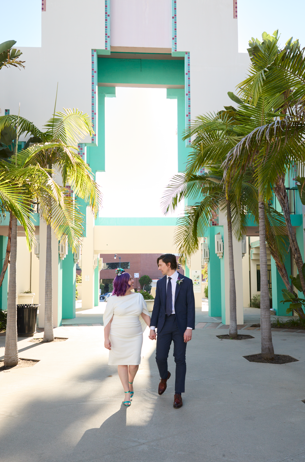 A couple in wedding attire walks hand-in-hand past tropical palm trees and a turquoise Art Deco building.