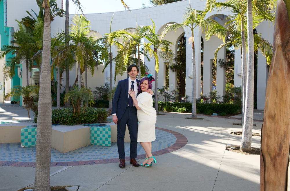 Couple posing together in a sunny courtyard with palm trees and white arched buildings in the background.