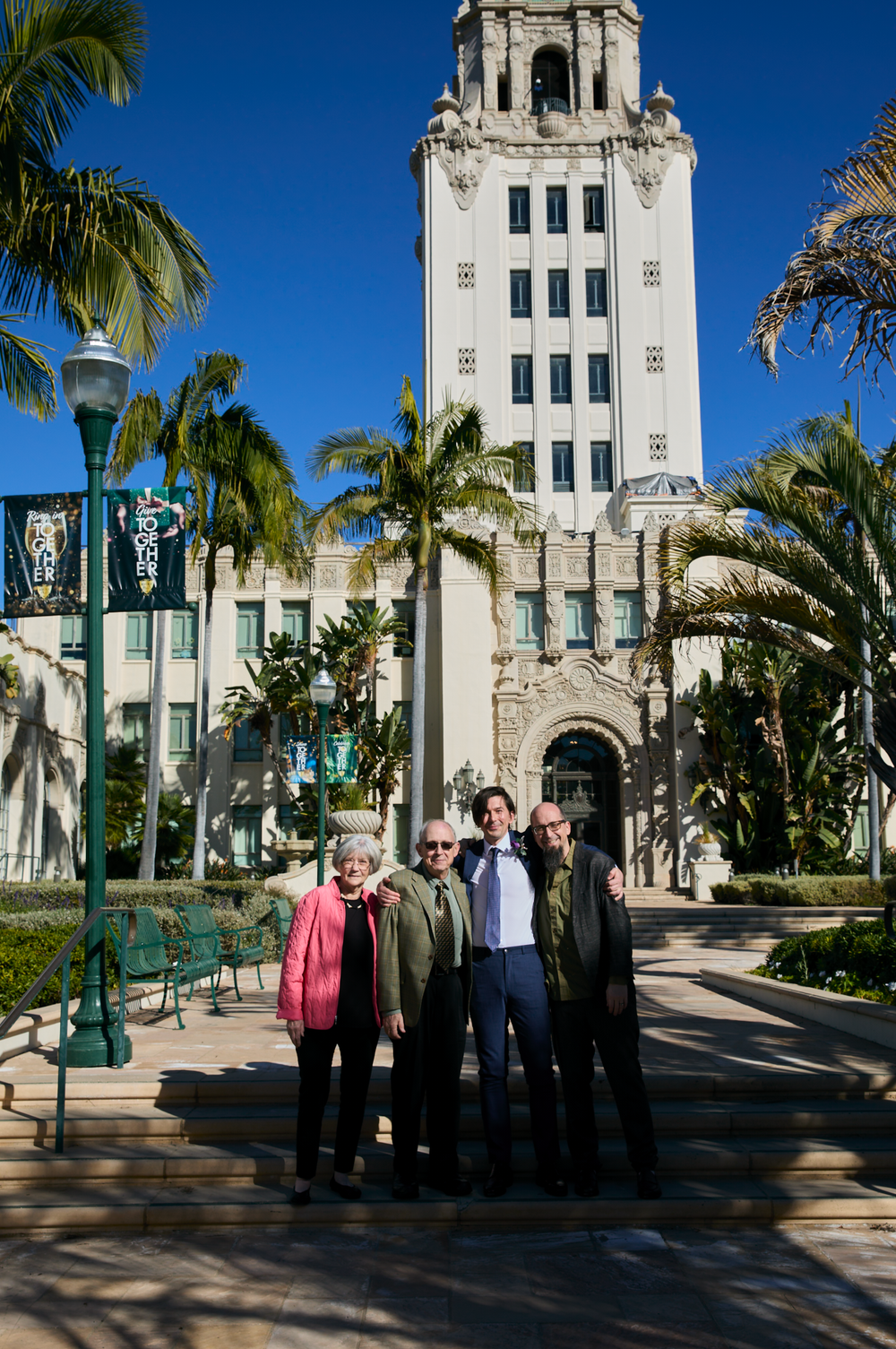 Four people pose in front of Beverly Hills City Hall, surrounded by palm trees under a clear blue sky.