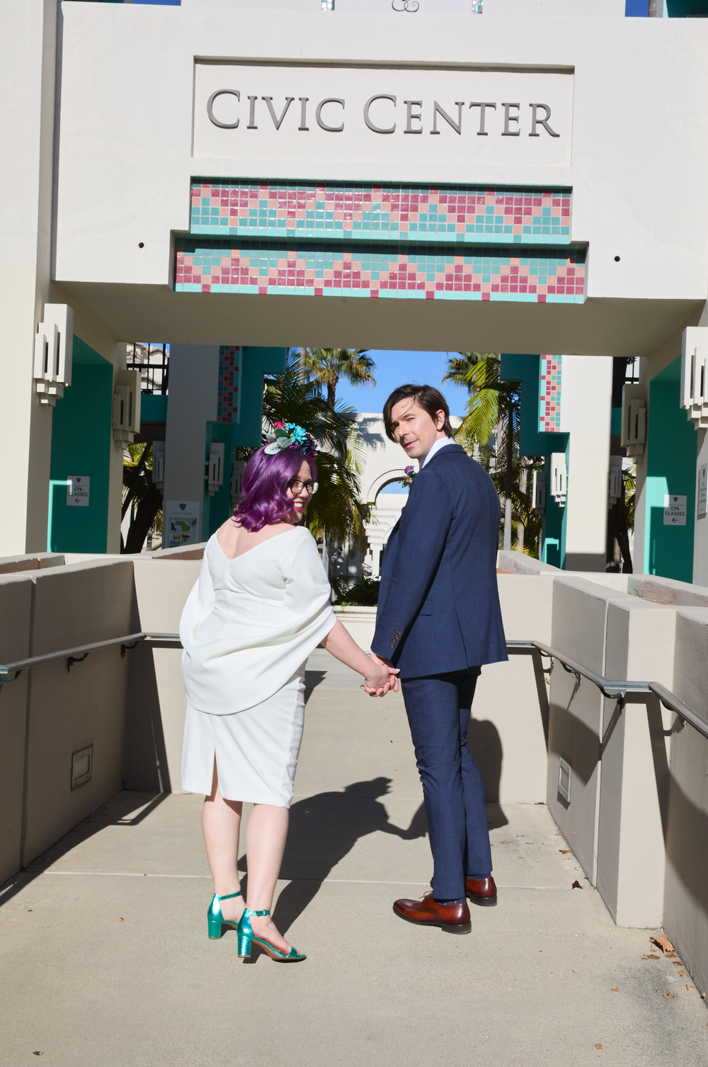 Couple holding hands in front of Civic Center, bride in white dress with purple hair, groom in navy suit.