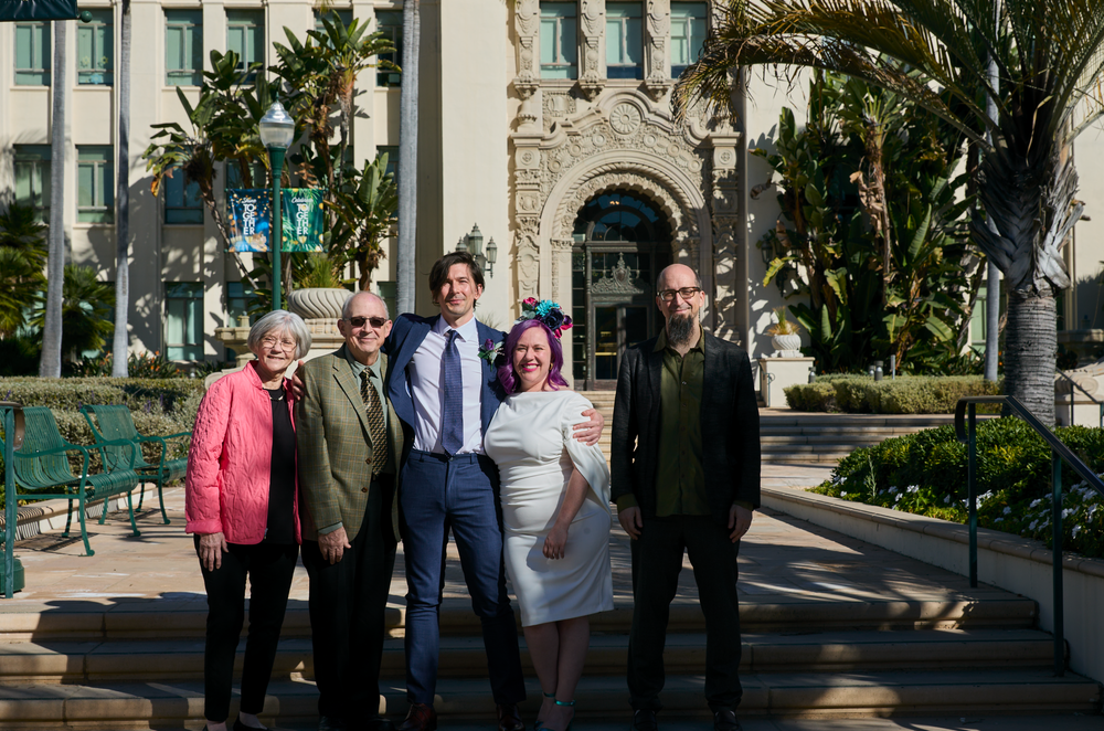 Five people posing together outdoors in front of an ornate building, smiling for a group photo on a sunny day.