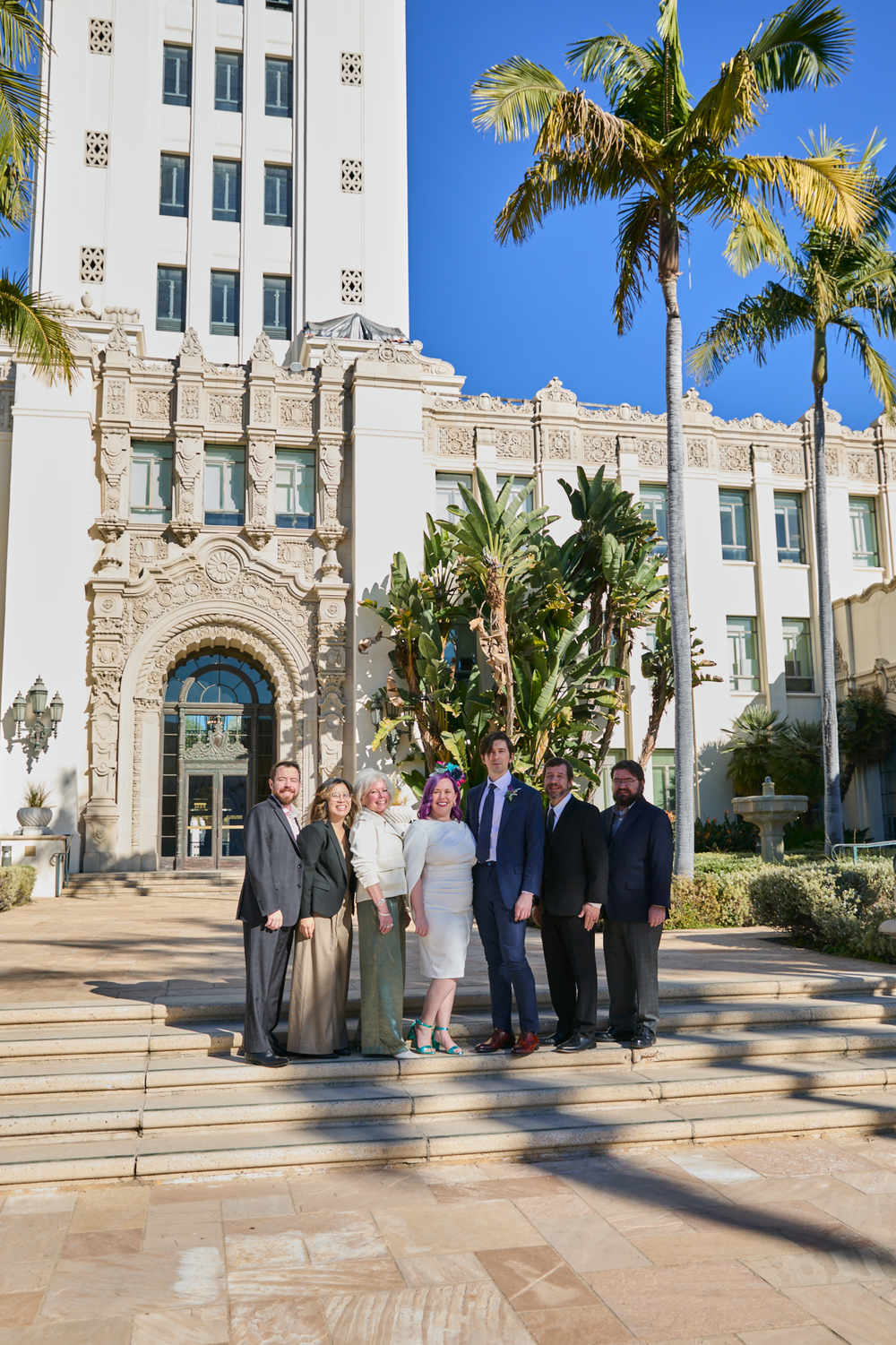 Group of seven professionals posing on steps outside ornate white government building with palm trees under blue sky.