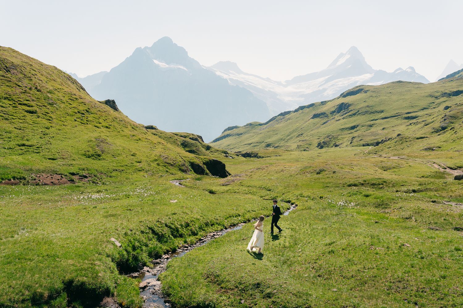 Couple in wedding attire walking through lush green alpine meadow with snow-capped mountain peaks in background.