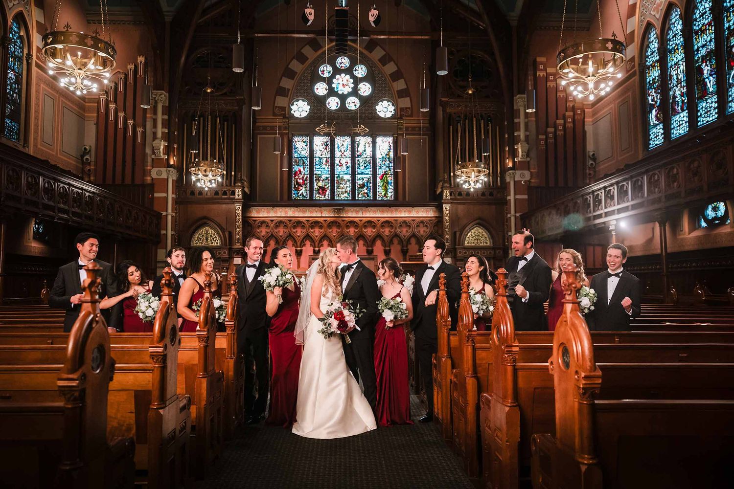 Large wedding party gathered in the aisle of Old South Church in Boston, bride and groom kissing surrounded by bridesmaids and groomsmen