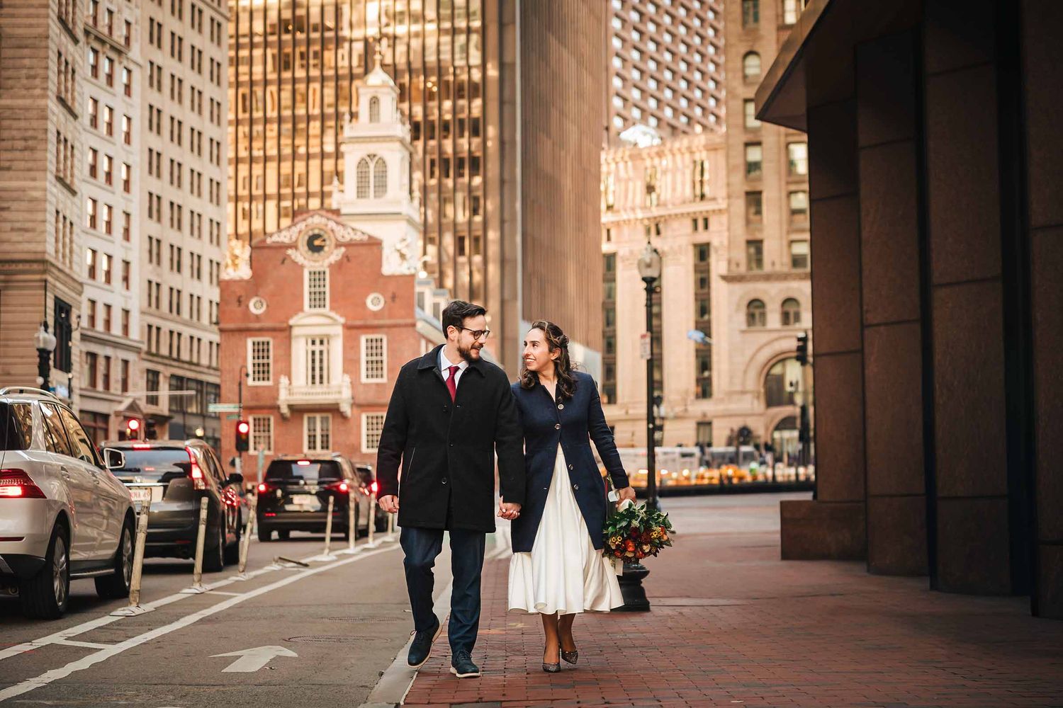 Couple smiling and holding each other on a Boston street near the Old State House, relaxed wedding portrait with fall bouquet