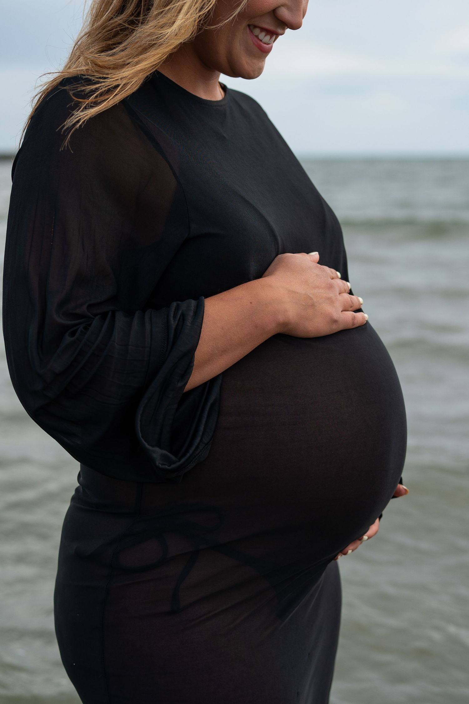 Smiling pregnant woman in black dress cradling belly by sea.