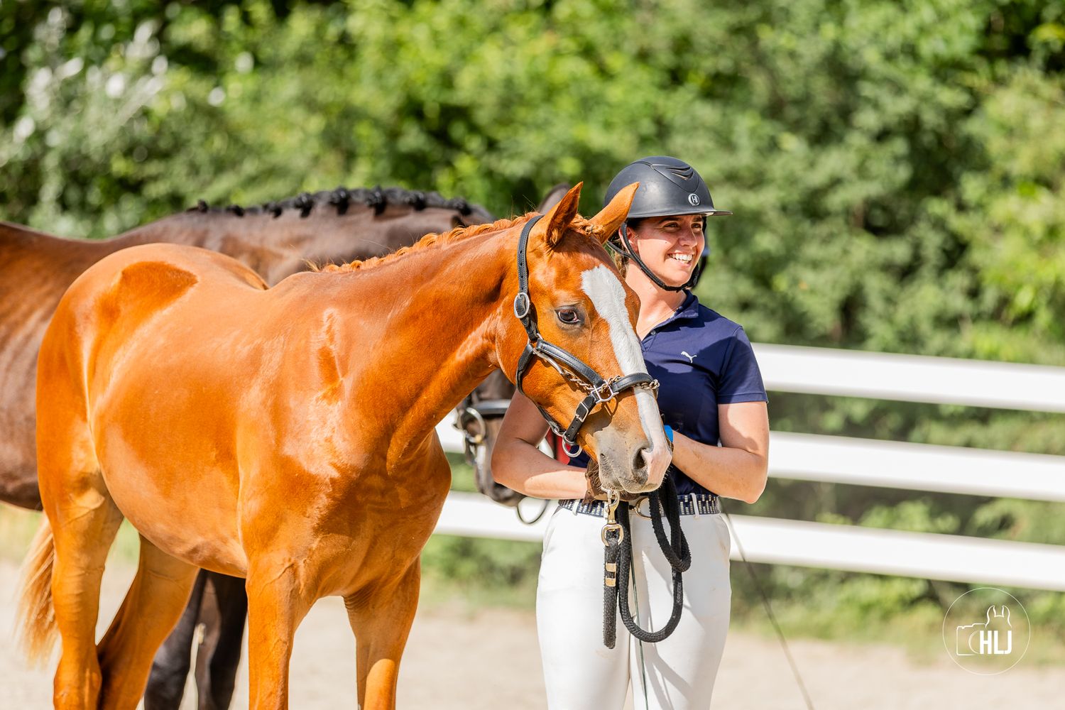 A female equestrian in a navy top and helmet stands beside a chestnut horse in an outdoor riding arena.