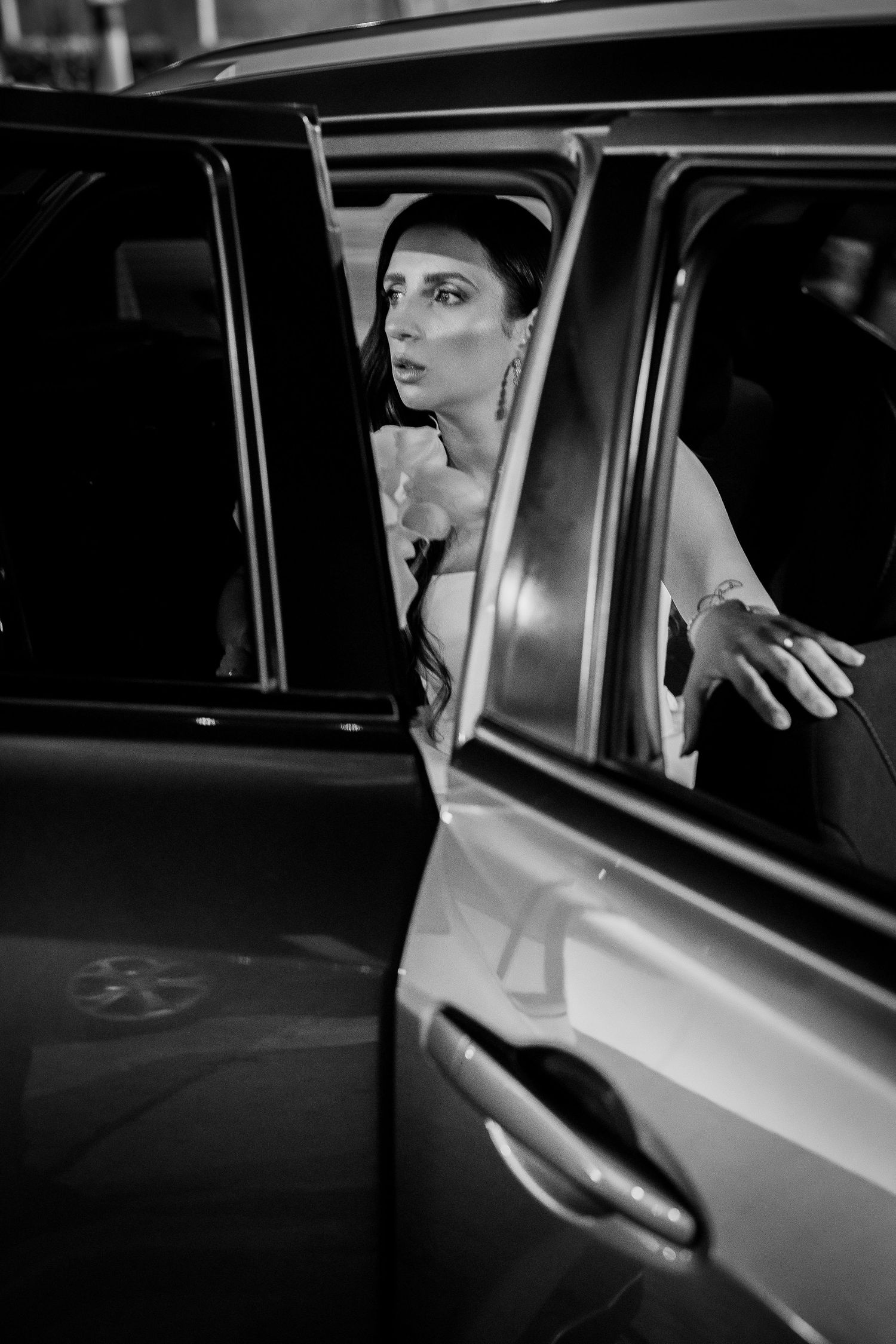 Black and white photo of an elegant bride stepping out of a luxury car, wearing a strapless gown and earrings.