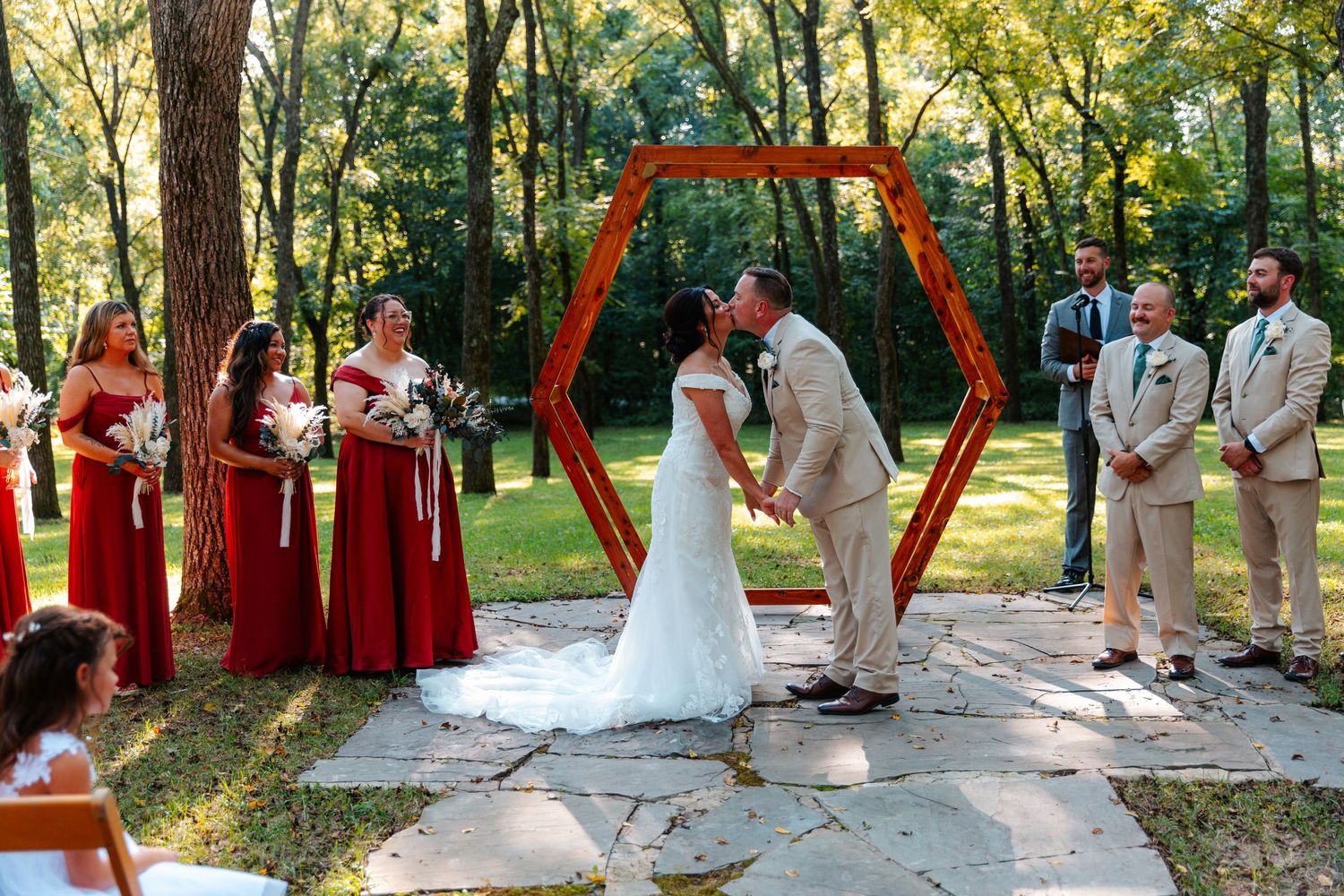 Bride and groom share first kiss under hexagonal wooden arch, flanked by red-gowned bridesmaids in outdoor woodland ceremony.