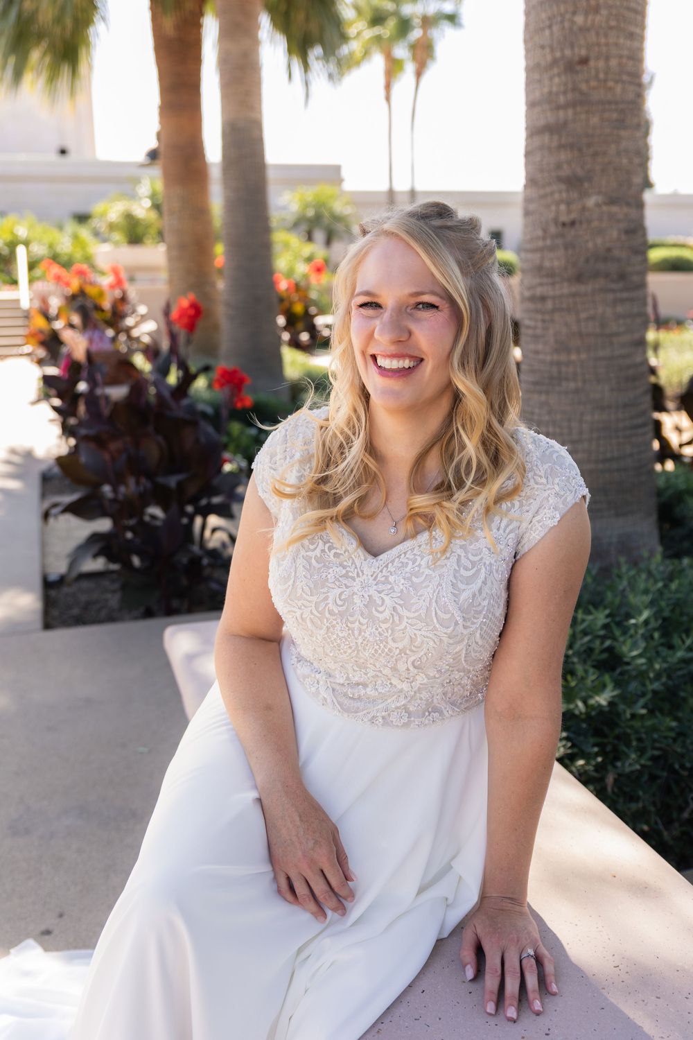 Smiling blonde bride in white lace wedding dress sitting outdoors near palm trees and tropical flowers on a sunny day.