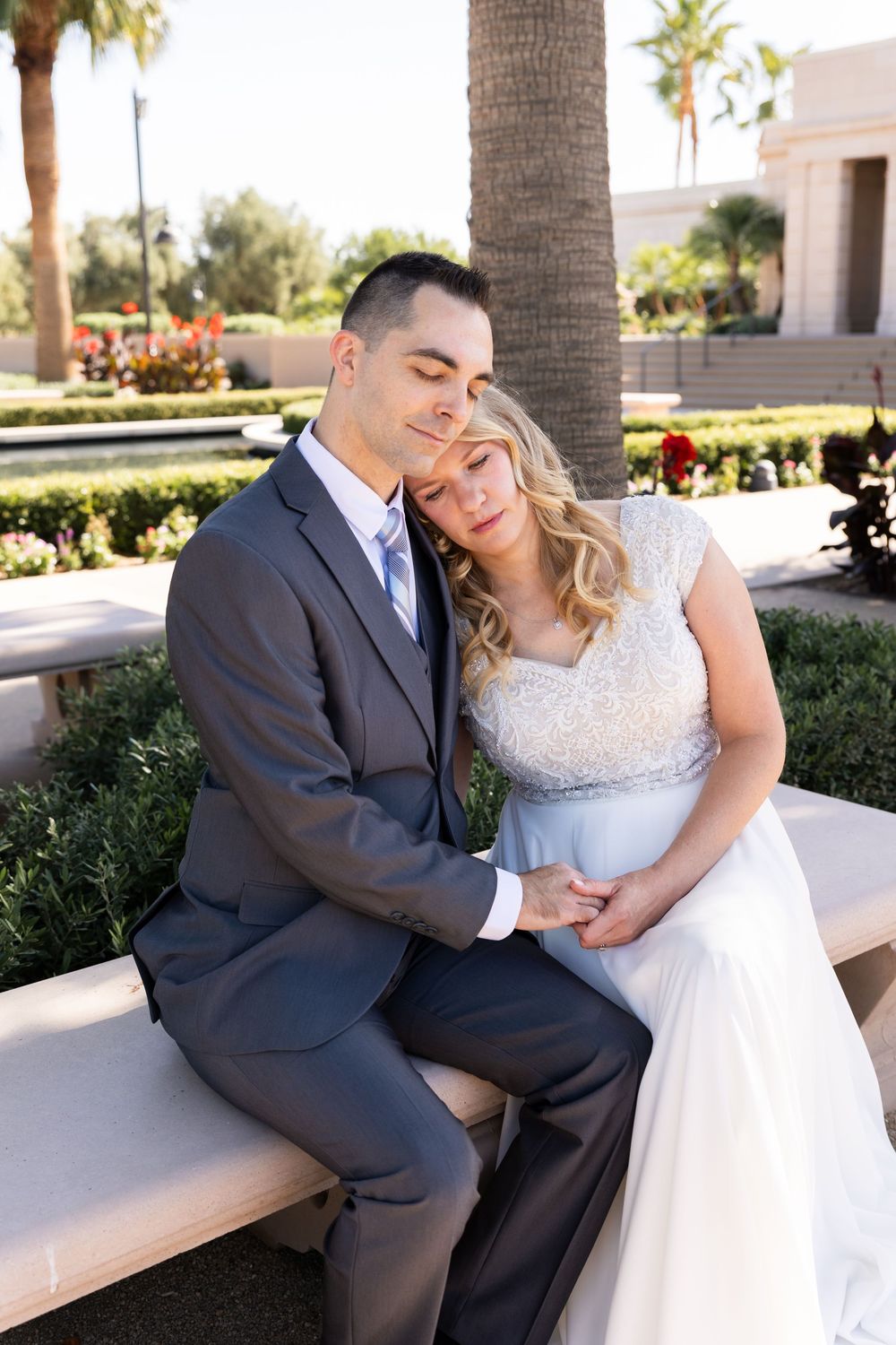 A bride and groom sit closely together on a stone bench outdoors, surrounded by lush greenery, sharing a tender moment.