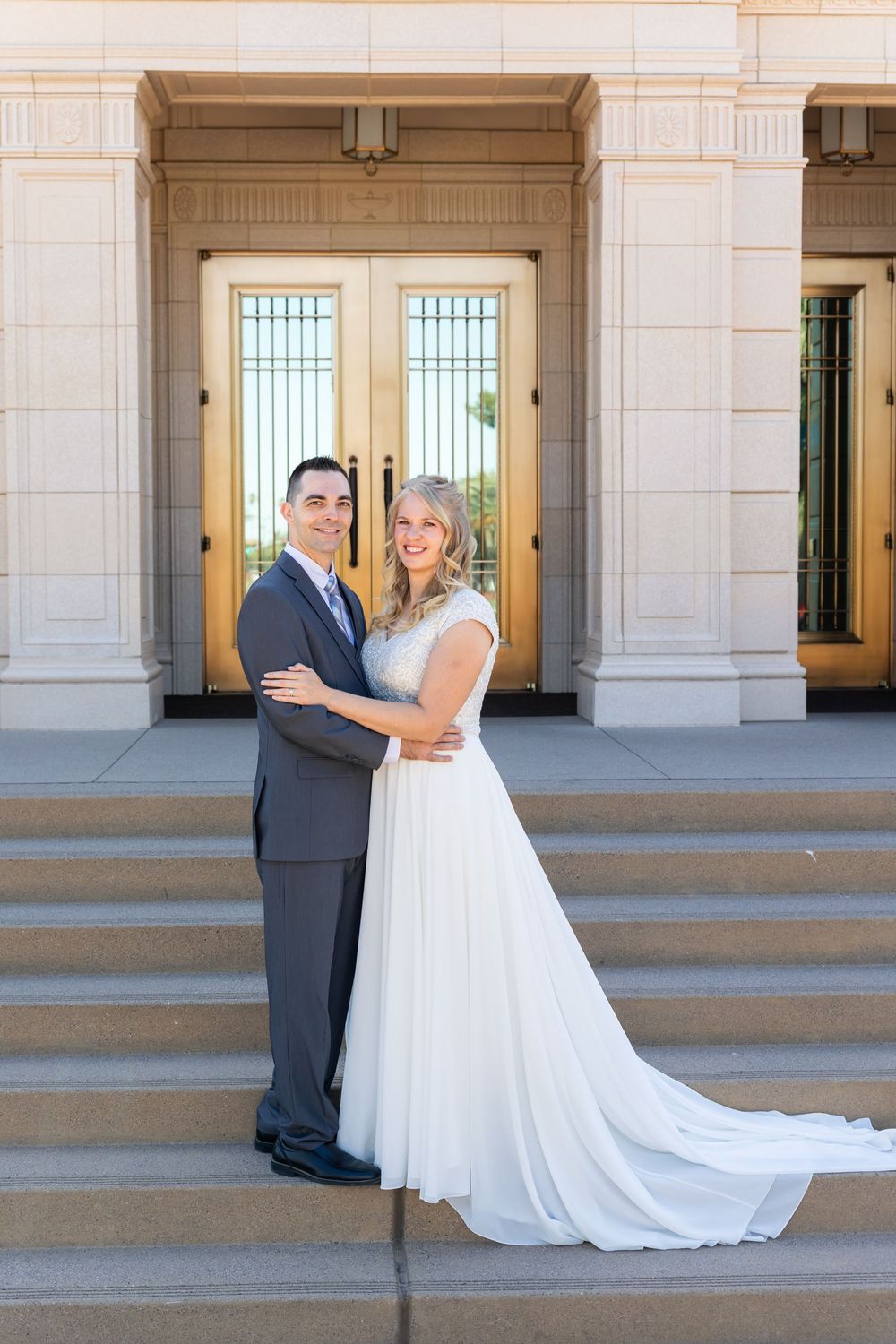 A smiling bride and groom pose on steps before elegant golden doors in a classic wedding portrait.