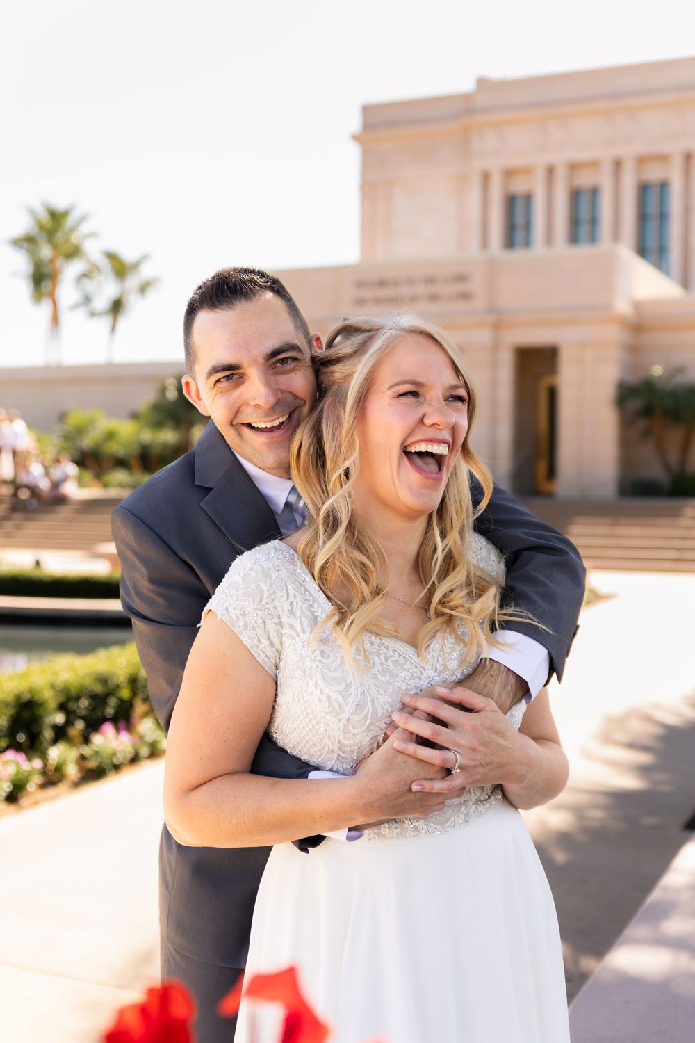 Joyful bride and groom laughing together outside a temple on their wedding day, bride in lace gown, groom in navy suit.