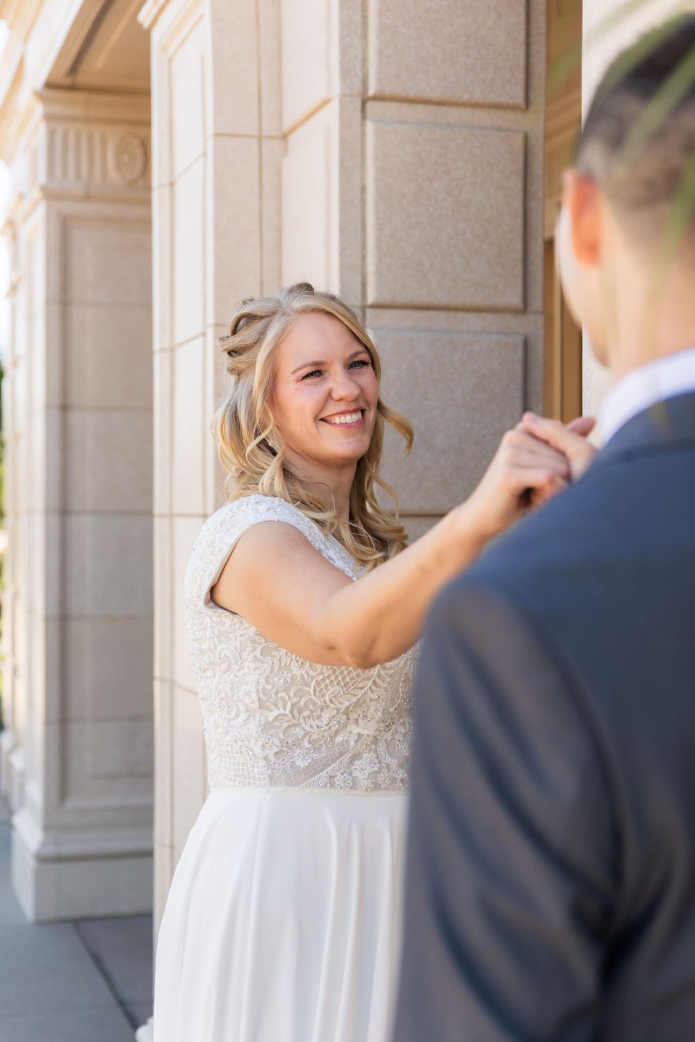 Smiling blonde bride in lace wedding dress holding grooms hand beside stone column outdoors.