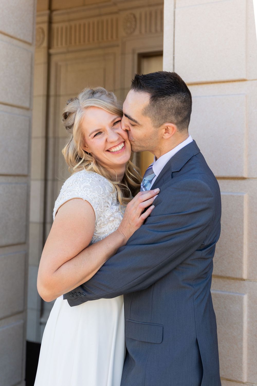 Groom in blue suit kissing smiling bride on cheek while embracing against stone building wall.