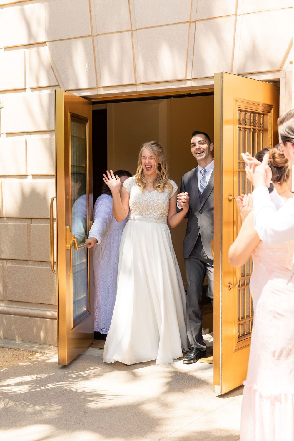 Joyful bride and groom exit through golden doors after wedding ceremony, greeted by cheering guests outside.