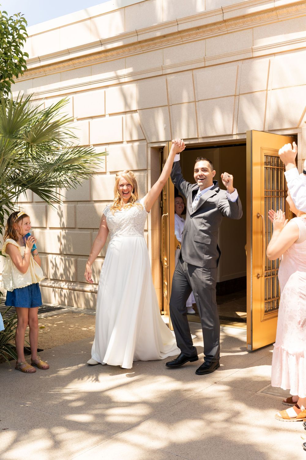 Joyful newlyweds exit wedding venue with raised hands as guests celebrate outside a grand building.