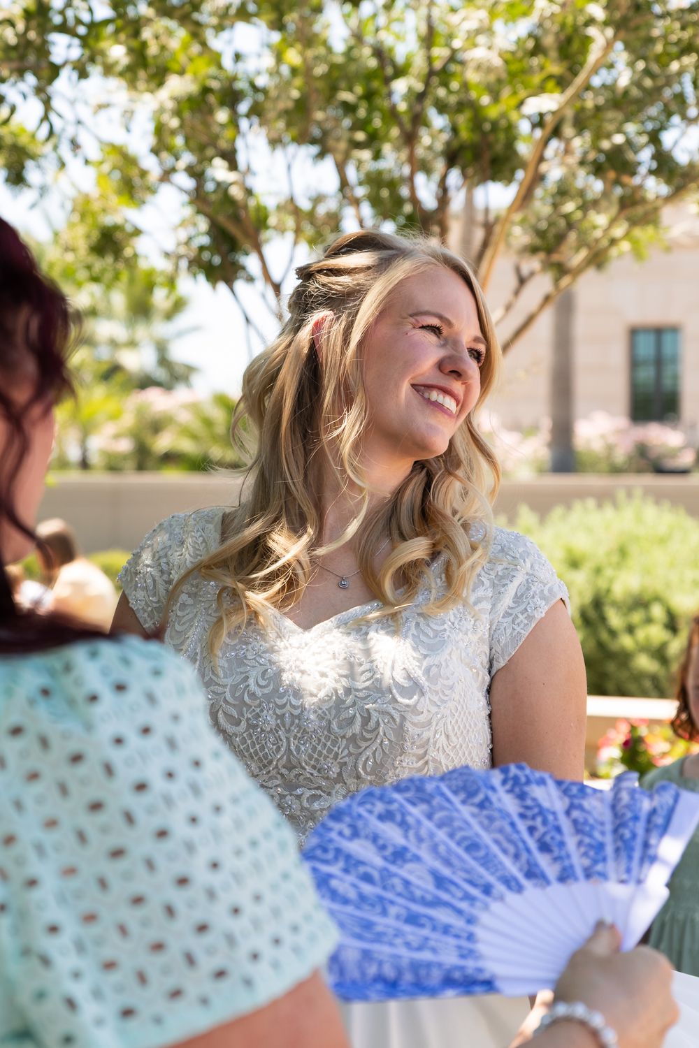 Happy blonde bride in lace wedding dress smiling outdoors while guest holds decorative blue fan.