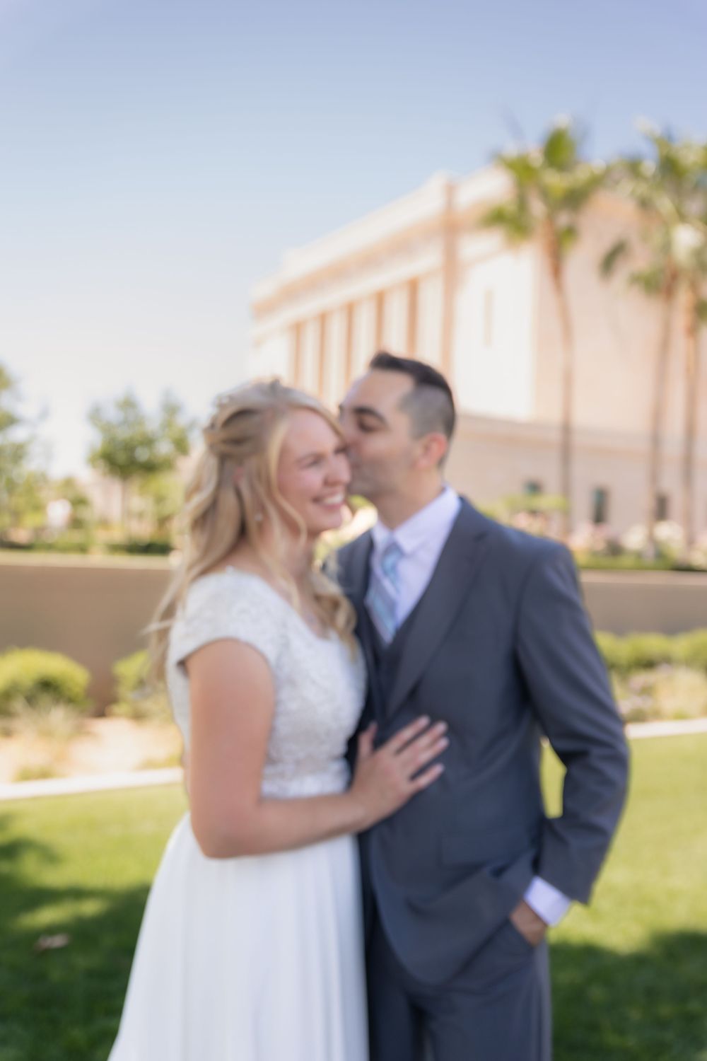 Bride and groom share a tender kiss outdoors near a white building with palm trees on their wedding day.