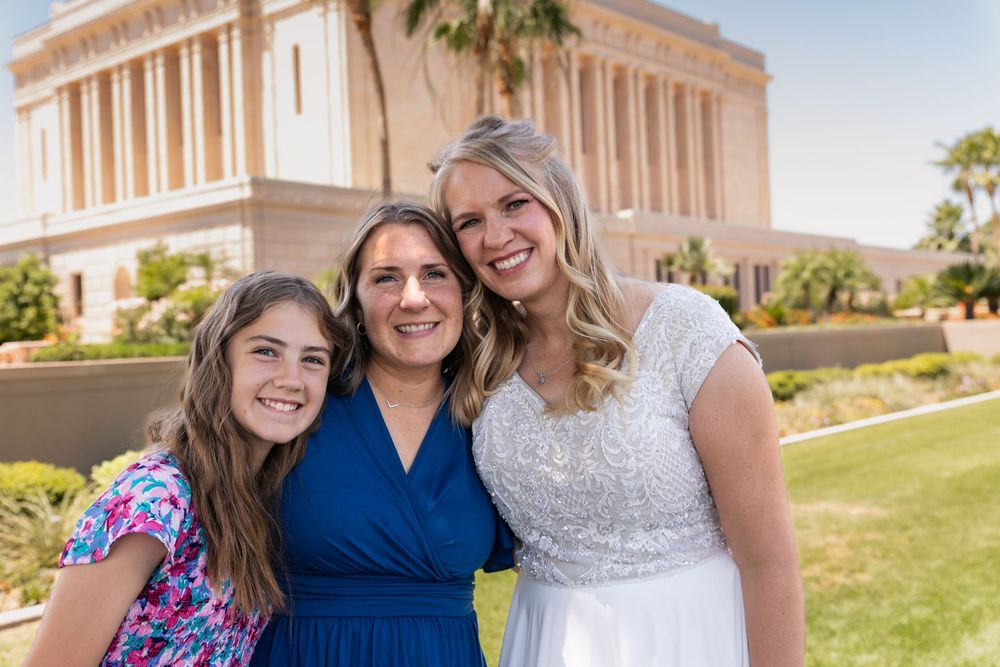 Three women smile together outdoors near a white temple, one wearing a wedding dress, on a sunny day.