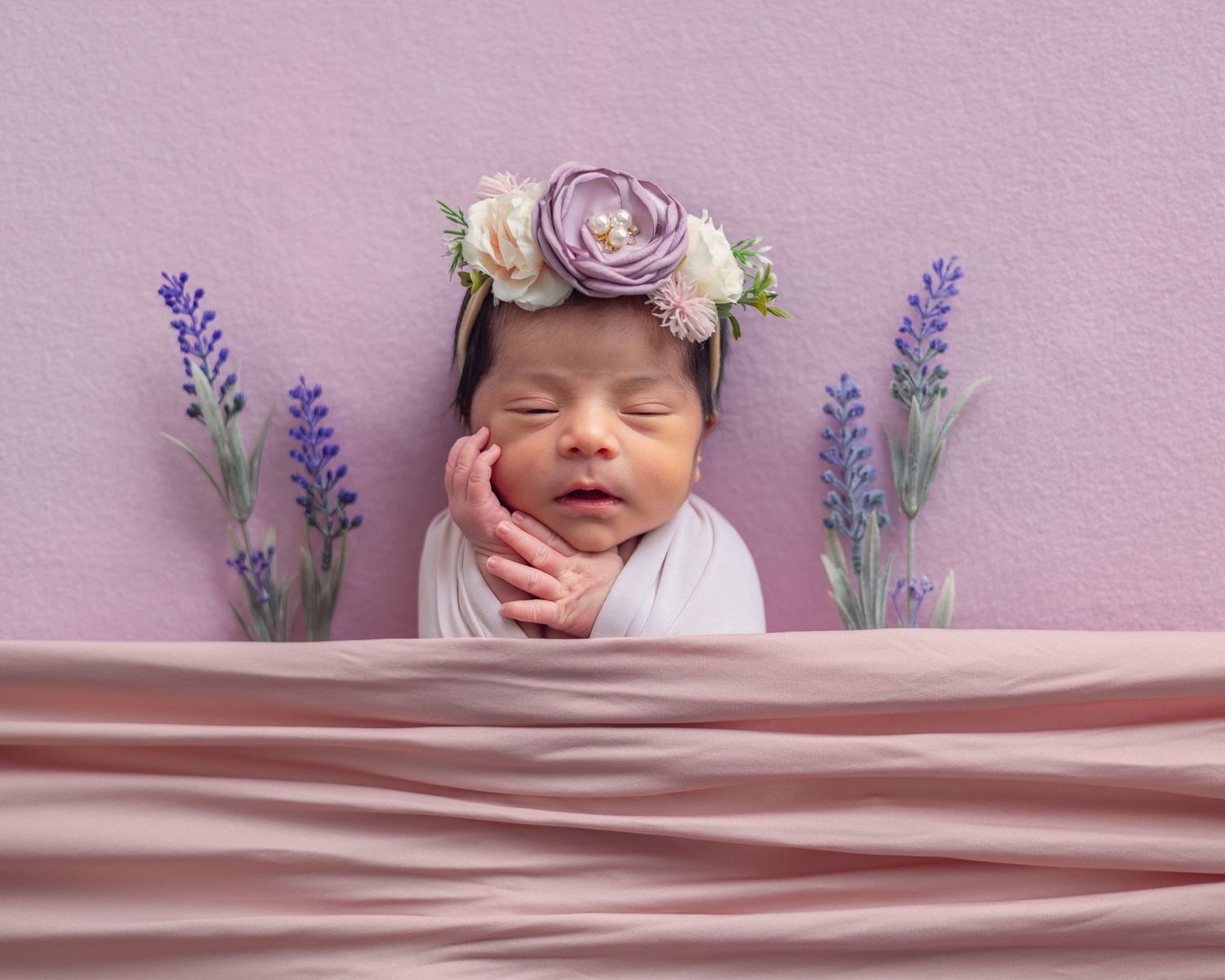 Sleeping newborn baby wrapped in pink with a floral crown, surrounded by lavender flowers on a soft pink background.