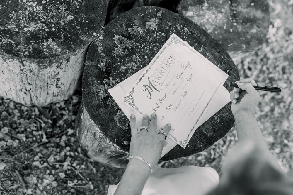 Close-up black and white photo of hands signing a marriage certificate outdoors on a rustic stone surface.