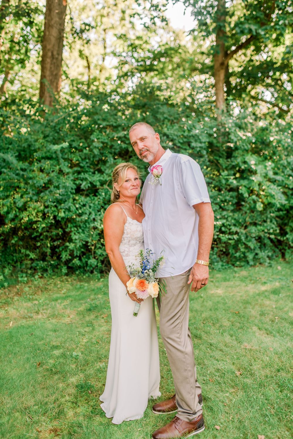 Bride and groom posing together outdoors on green lawn, bride holding colorful bouquet in white gown, groom in light shirt.