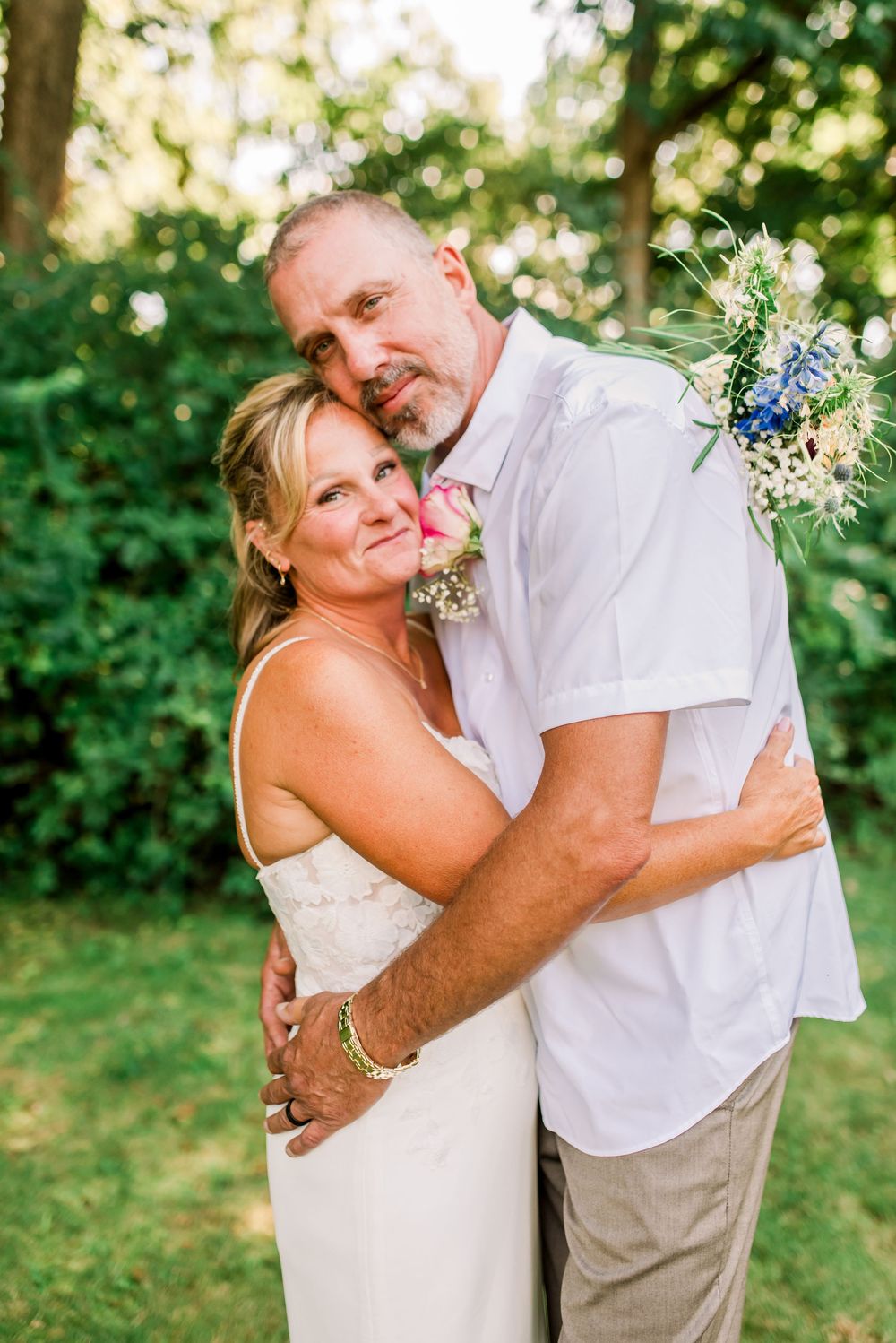 A couple in wedding attire embraces outdoors, the bride in white holding a bouquet, surrounded by lush greenery.
