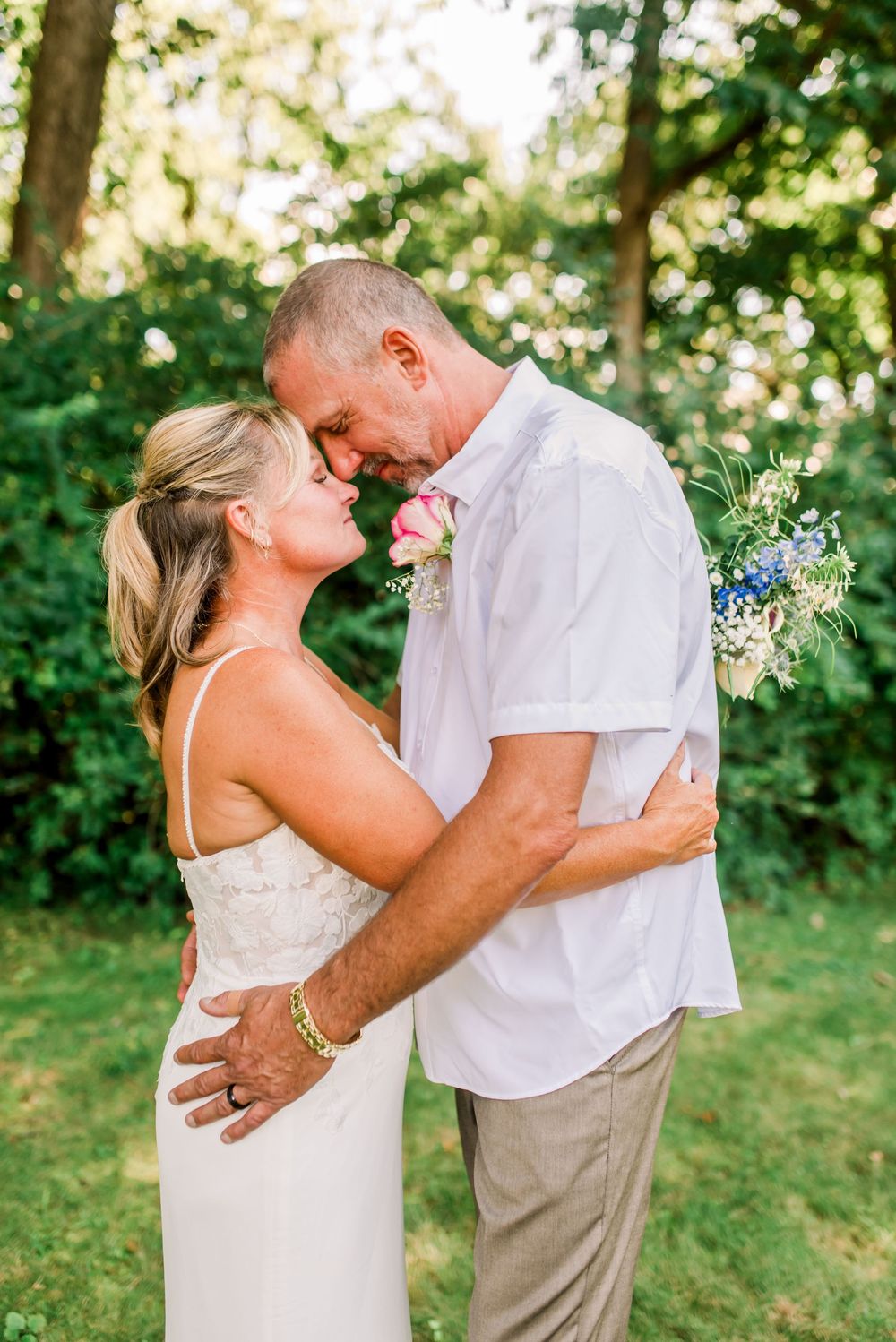 Couple in wedding attire embracing and smiling outdoors, groom in white shirt with boutonniere, bride in white dress.