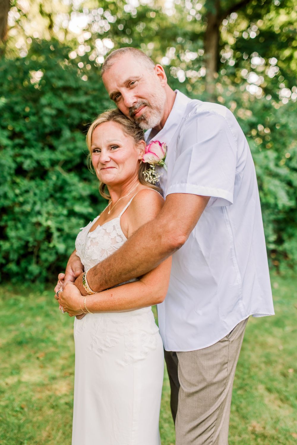 A couple in wedding attire embraces outdoors, the bride in white dress and groom in light blue shirt with boutonniere.