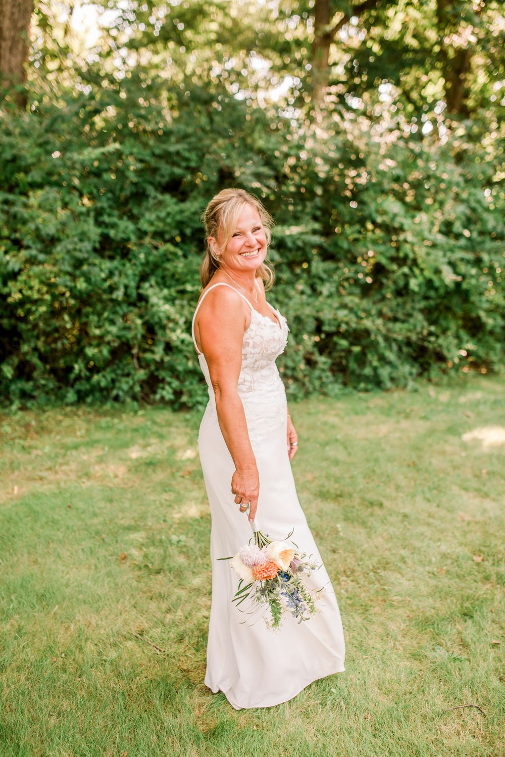 Smiling bride in white sleeveless gown holding pastel bouquet standing on green lawn with lush trees backdrop.