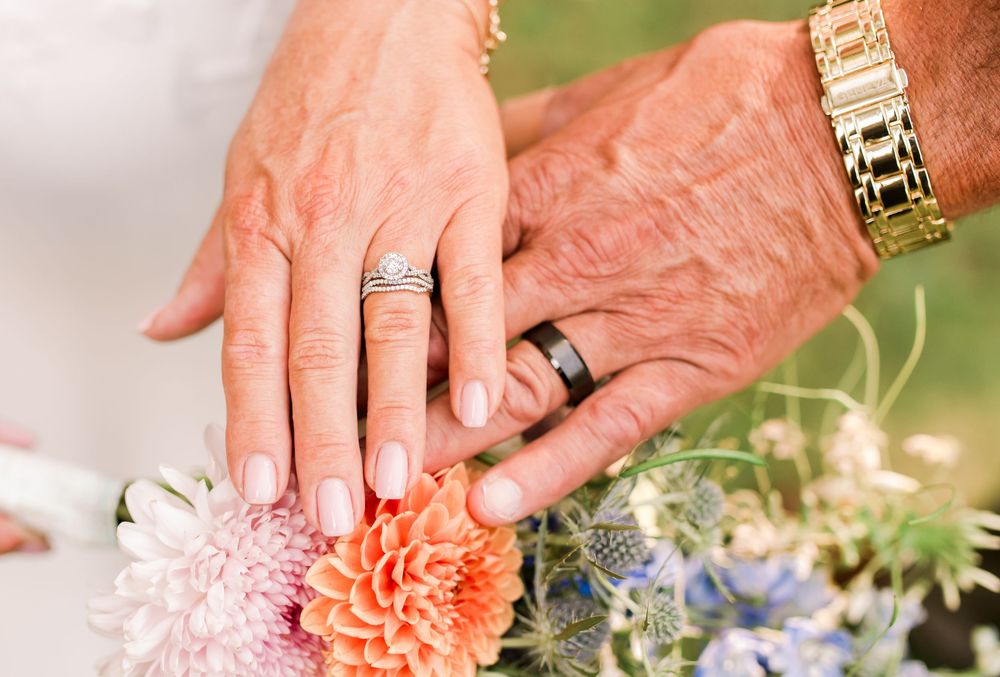 Two elderly hands displaying wedding rings rest on a colorful bridal bouquet with dahlias and blue flowers.