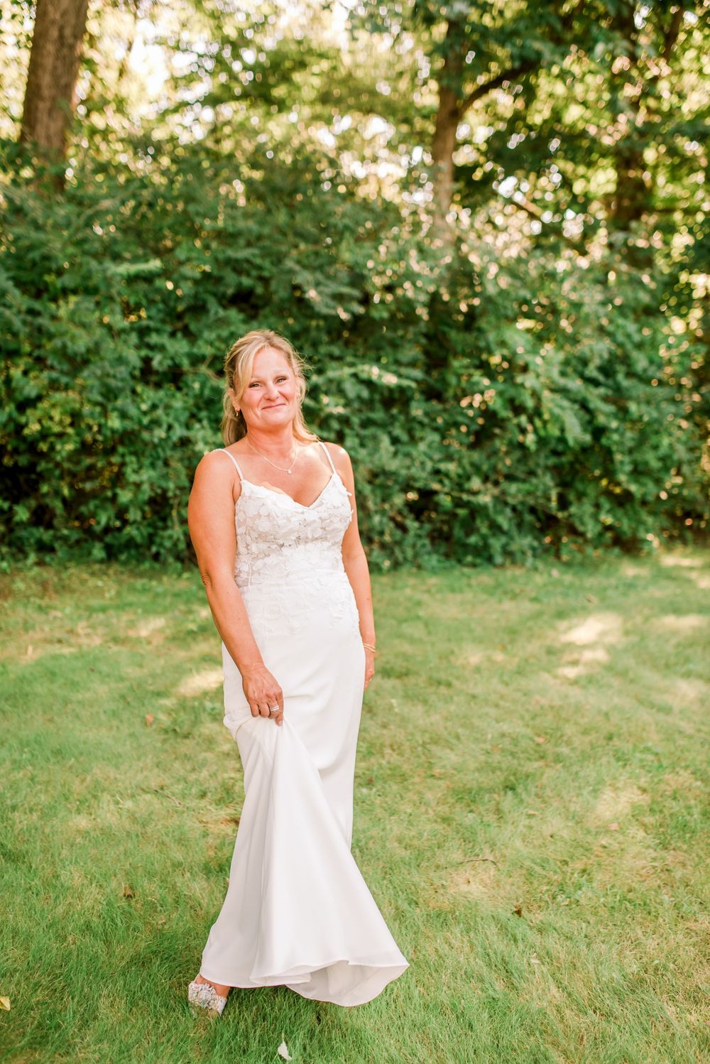 Bride in white spaghetti strap wedding gown with lace bodice smiling outdoors on green lawn with trees.