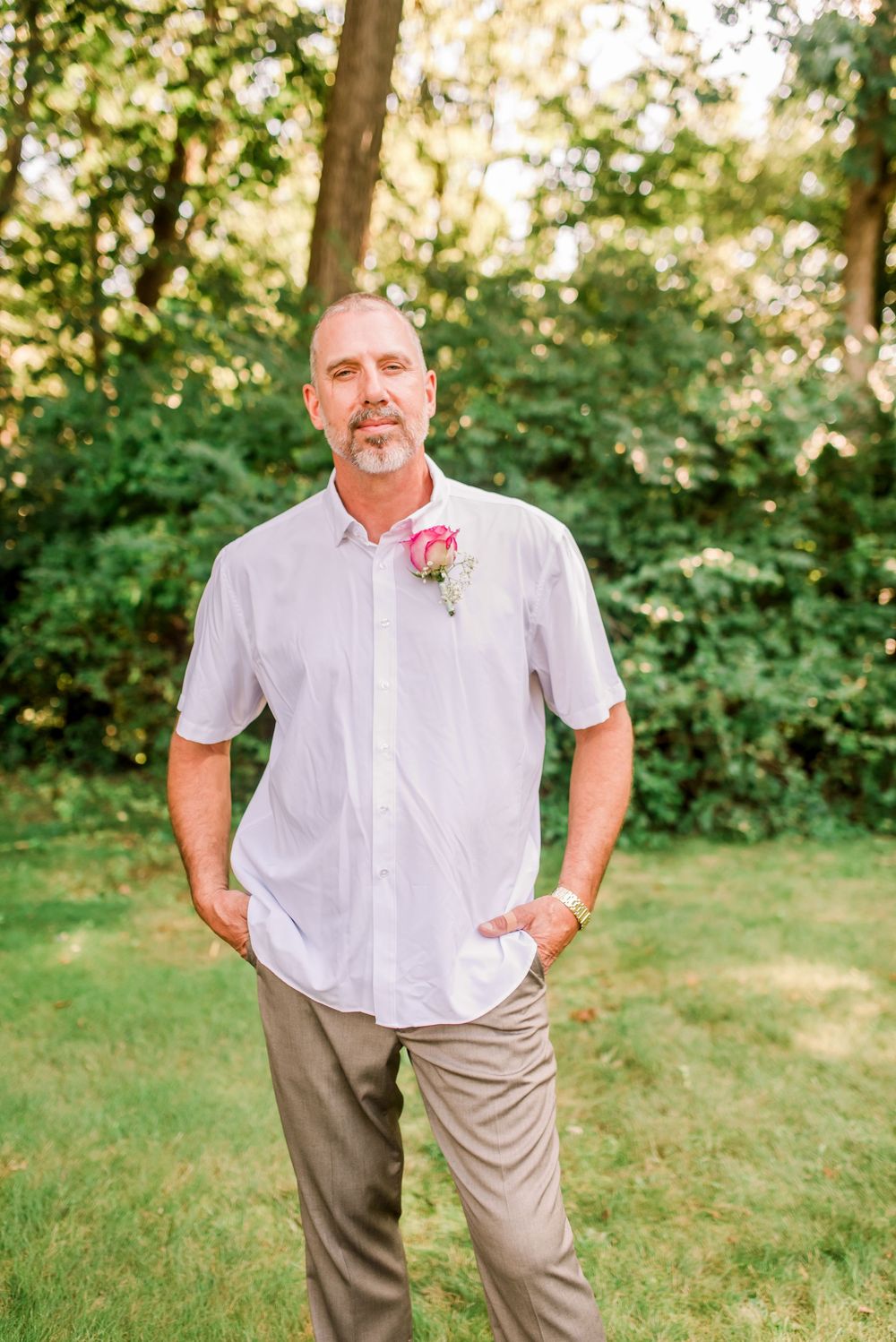 Groom in white short-sleeve shirt with pink boutonniere standing confidently on green lawn with trees behind.