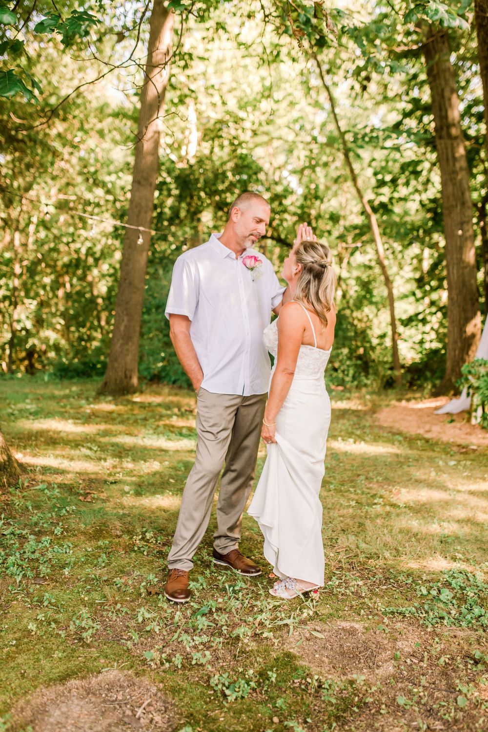Bride and groom share a tender moment outdoors in a sunlit forest setting during their wedding day.