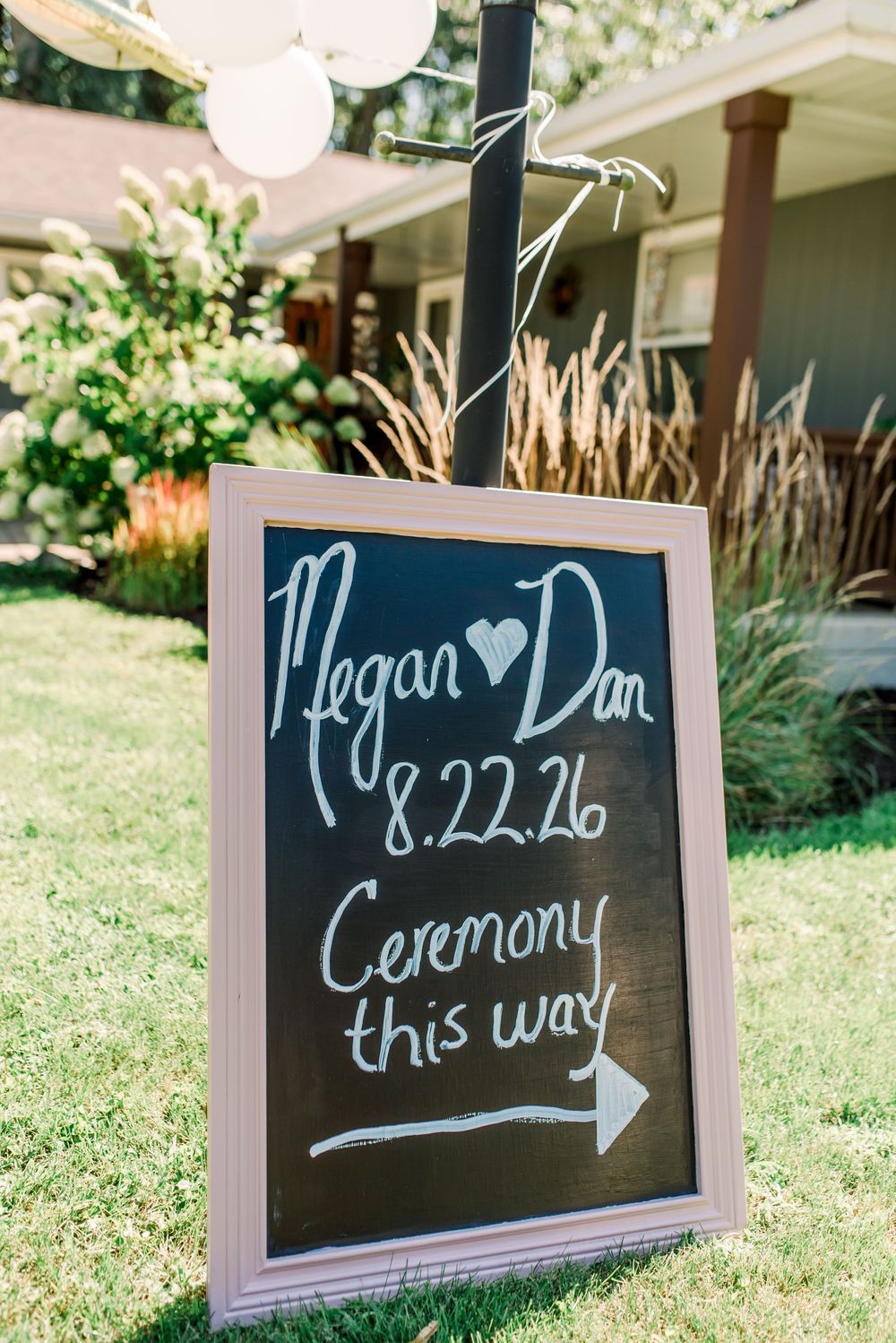 Chalkboard wedding sign reading Megan and Dan 8.22.26 Ceremony This Way with arrow, displayed on green lawn.