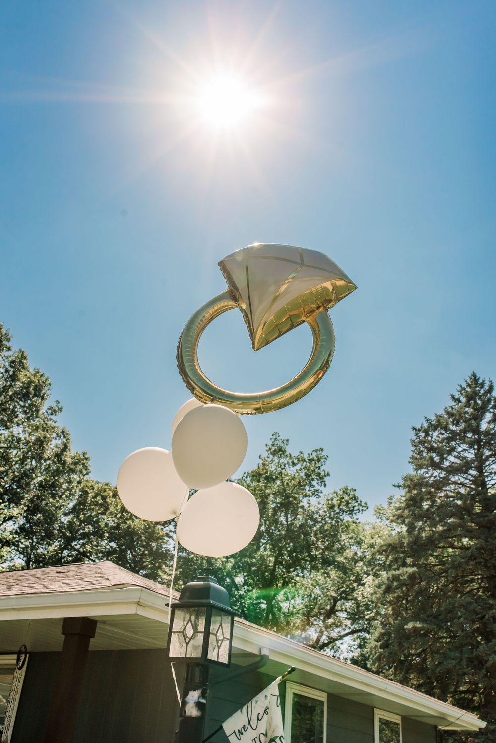 Diamond ring balloon floating in sunny blue sky with white balloons below, trees and house roof in background.