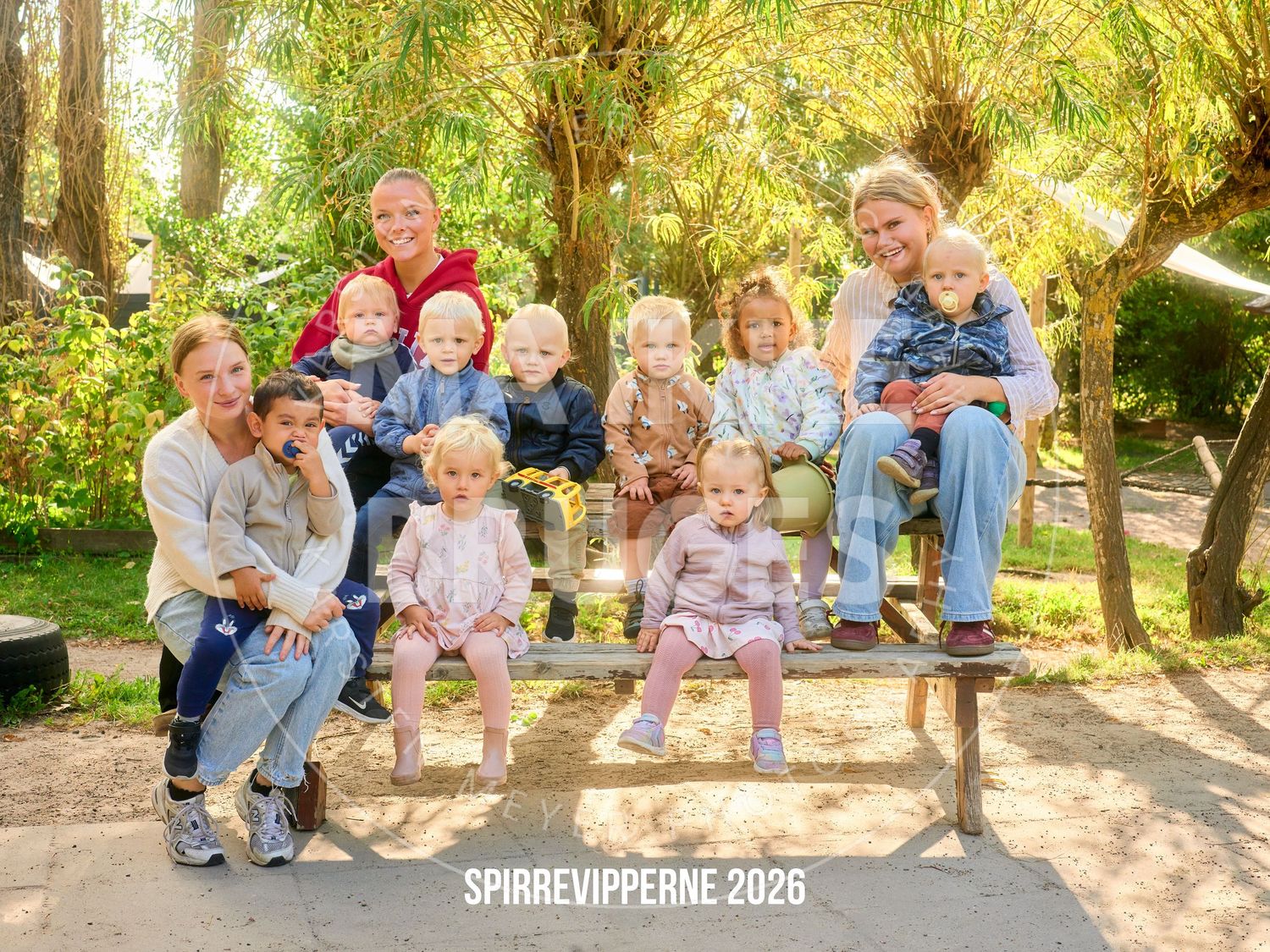 Family group photo with adults and young children on a bench in a lush green park, labeled Spirrevipperne 2026.