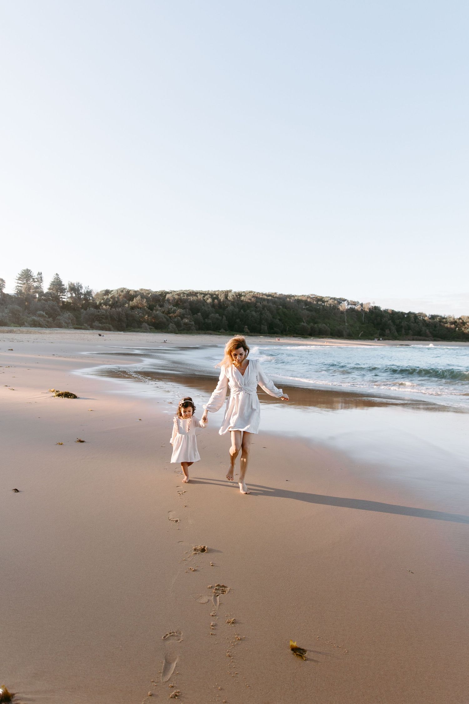 A mother and young daughter in matching white dresses walk hand-in-hand along a sandy beach at sunset.