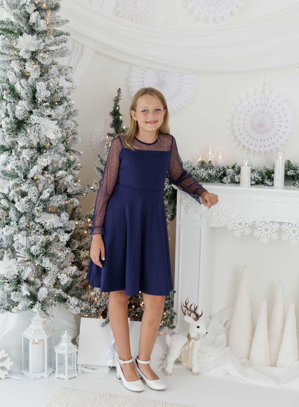 Girl in navy blue dress with sheer sleeves poses by flocked Christmas tree and white fireplace mantel.