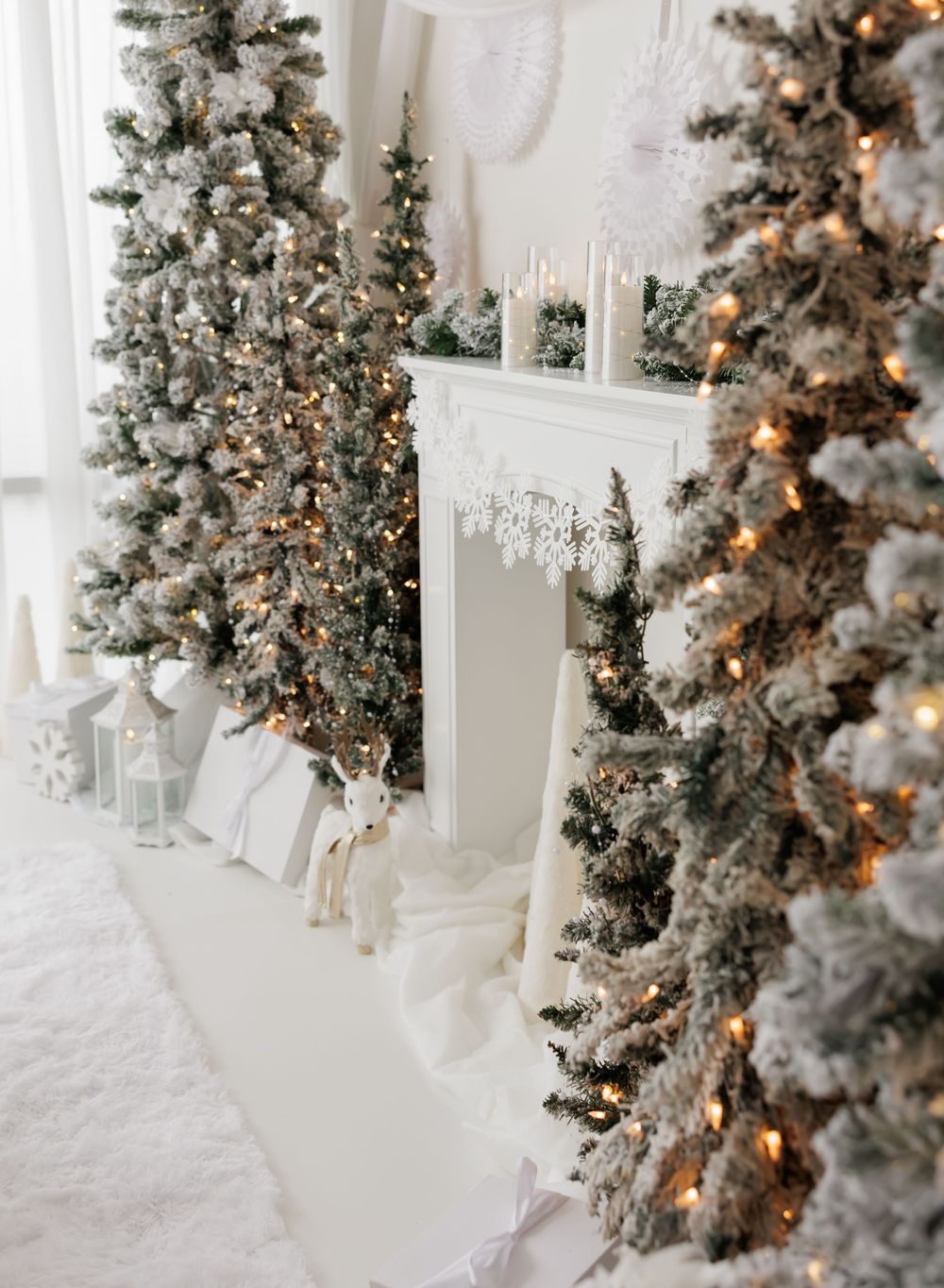 Elegant white Christmas room with snow-flocked trees, warm fairy lights, and a decorated fireplace mantel.