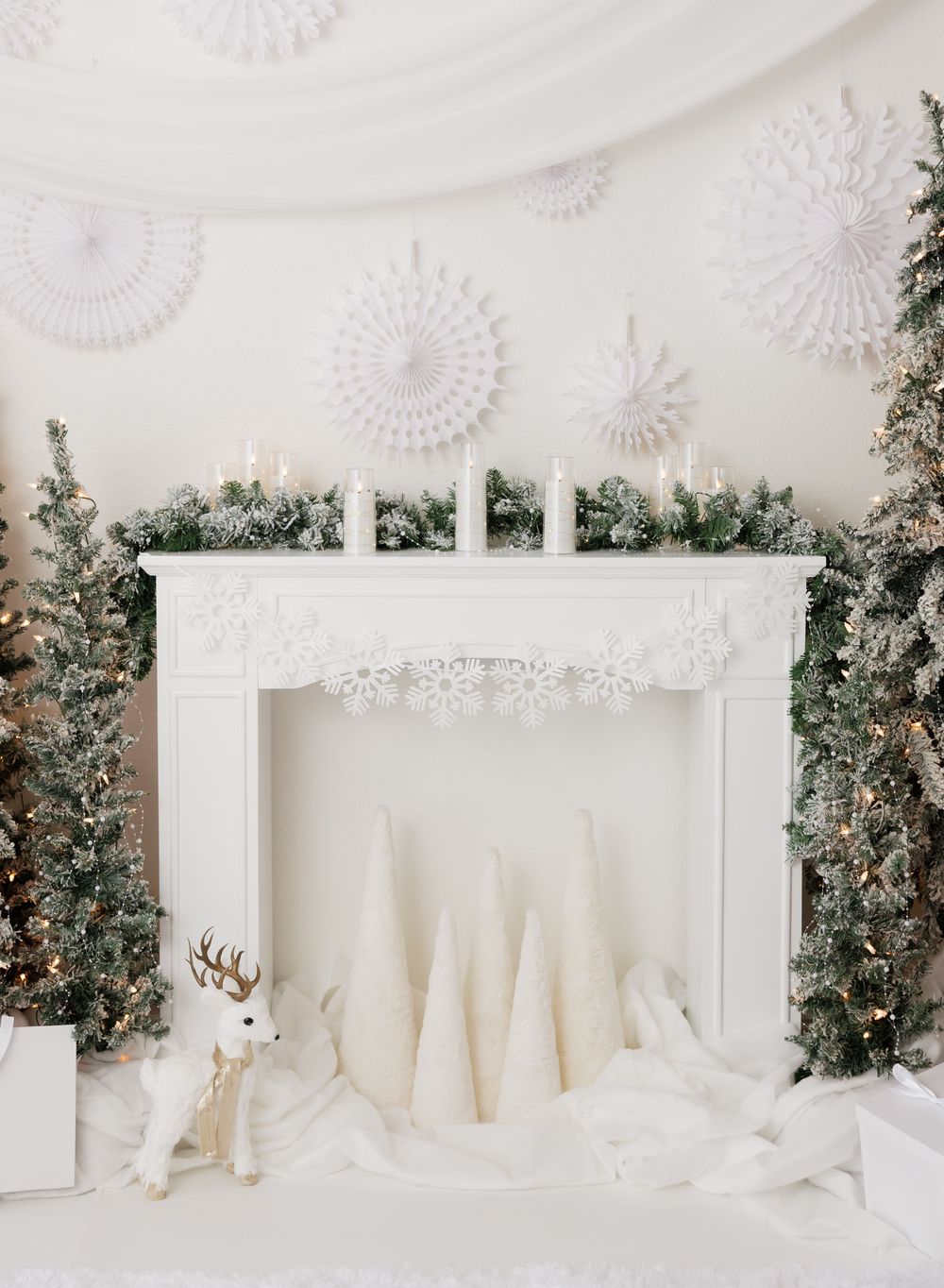 White Christmas fireplace decor with snowy trees, paper snowflakes, candles, garland, and a reindeer figurine.