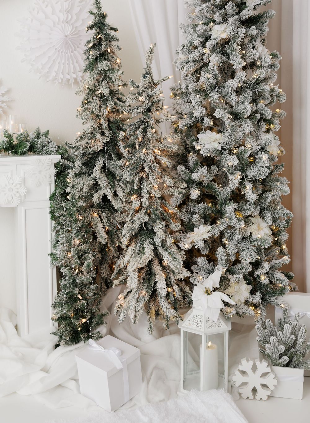 Snow-flocked Christmas trees with warm fairy lights, white gifts, and a lantern in a festive winter white holiday decor setting.