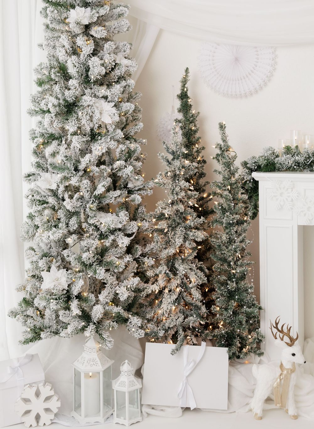 Snow-flocked Christmas trees with warm lights, white lanterns, gifts, and reindeer figurine in elegant holiday decor.