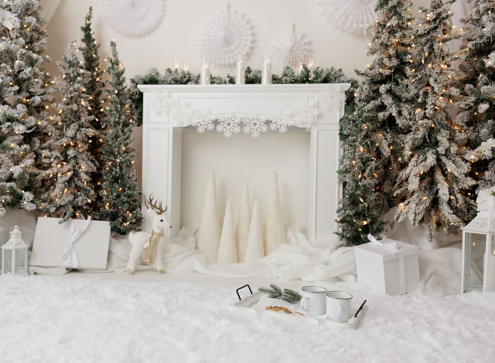 White Christmas fireplace scene with snow-flocked trees, reindeer figurine, wrapped gifts, and fairy lights on a white fur rug.