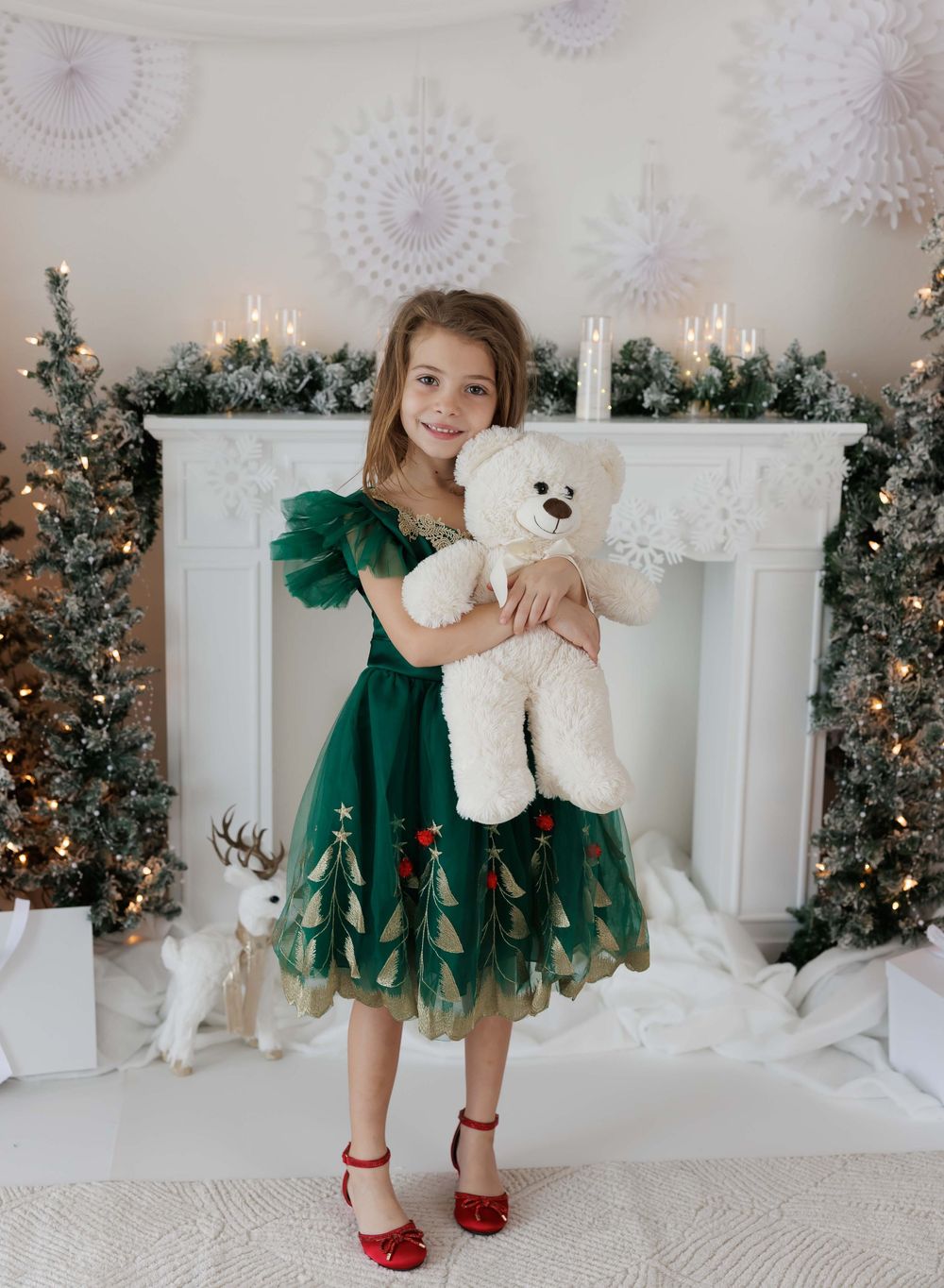 Girl in green Christmas tree tulle dress holding white teddy bear in front of decorated holiday fireplace.