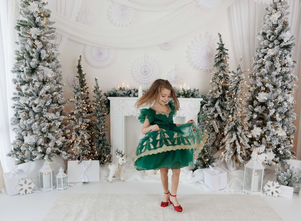 Young girl in green dress twirling among snow-flocked Christmas trees in a white holiday decorated room.