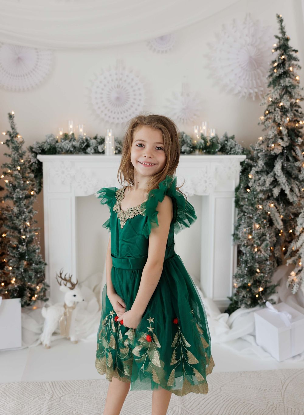 Girl in green Christmas tree dress poses before festive fireplace with snow-flocked trees and holiday decor.