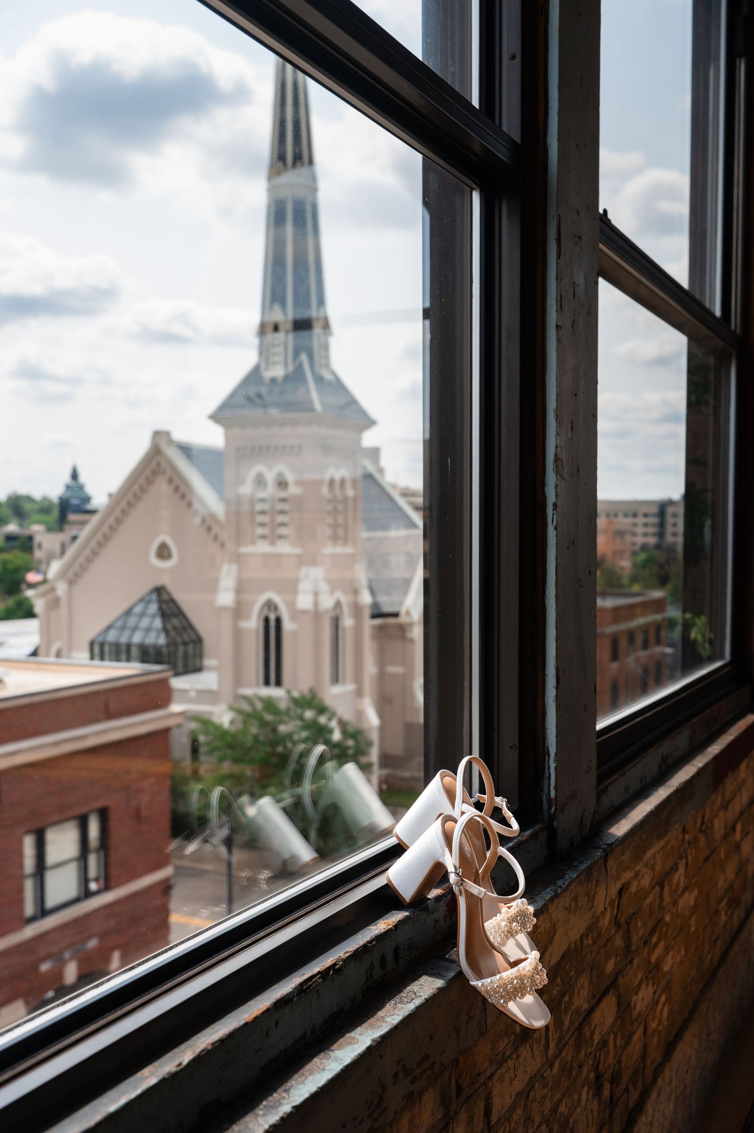 Bridal heels displayed on a window sill with a historic church steeple and brick buildings in the background.